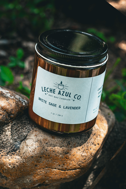 Close-up of an artisanal 7 oz Leche Azul Co. soy wax candle in the White Sage & Lavender scent. The candle is housed in a minimalist amber glass jar with a textured black lid and features a clean white product label. It is styled outdoors, resting on a textured, sunlit stone surface with natural soil and soft greenery blurred in the background.