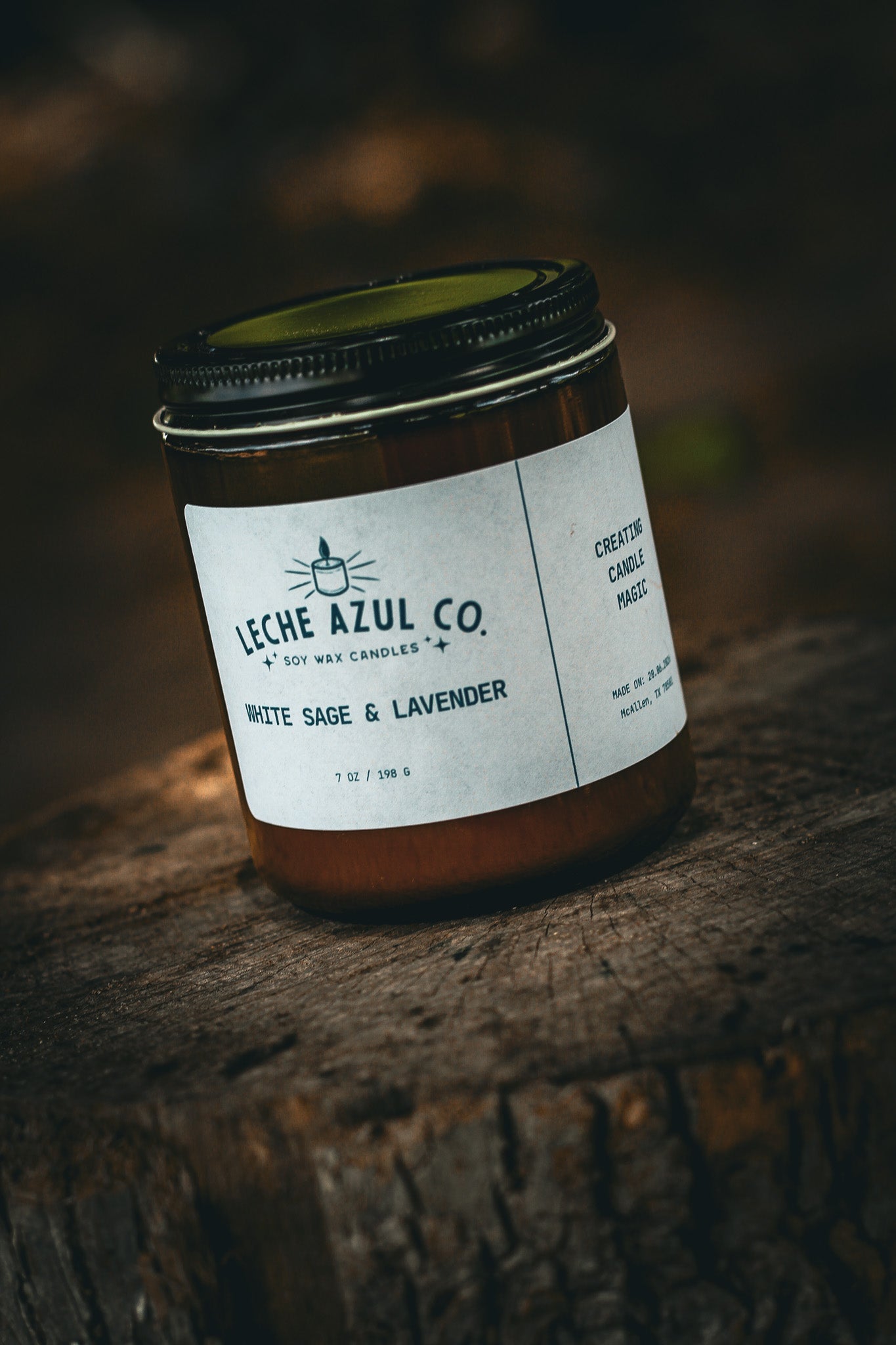 A close-up, angled shot of an artisanal 7 oz Leche Azul Co. soy wax candle in the White Sage & Lavender scent. The candle is housed in a minimalist amber glass jar with a black lid and a clean white product label reading "White Sage & Lavender" and "Creating Candle Magic". It is styled outdoors, resting on a textured, weathered tree stump under dramatic, natural lighting with a dark, earthy background.