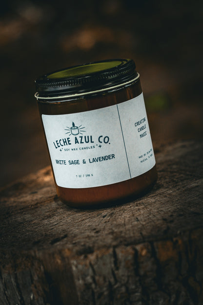 A close-up, angled shot of an artisanal 7 oz Leche Azul Co. soy wax candle in the White Sage & Lavender scent. The candle is housed in a minimalist amber glass jar with a black lid and a clean white product label reading "White Sage & Lavender" and "Creating Candle Magic". It is styled outdoors, resting on a textured, weathered tree stump under dramatic, natural lighting with a dark, earthy background.