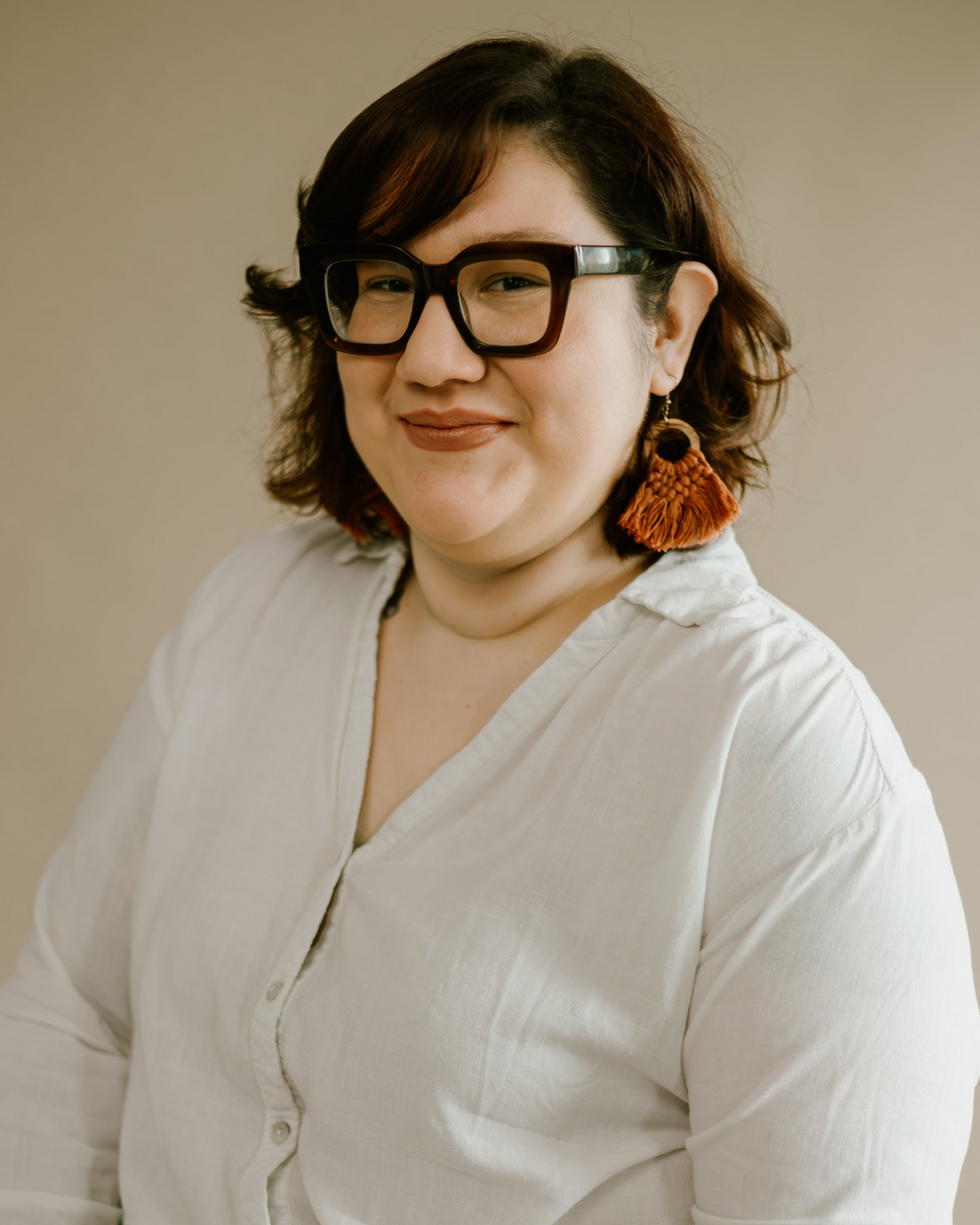 A professional studio headshot of a smiling woman with shoulder-length dark hair and auburn highlights. She is wearing thick-rimmed, dark square glasses, a white button-down collared linen shirt, and large, textured orange-brown macrame fringe statement earrings. She stands posed against a neutral, warm beige backdrop in a soft, bright lighting setting.